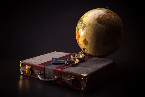 Still life on a dark background of an old fashioned globe and suitcase with a passport and a brass compass.