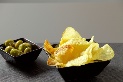 Two small servings of potato chips and green olives on black plates on top of a table.
