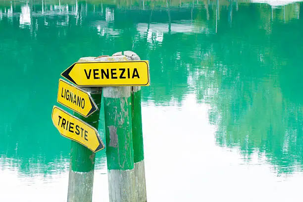 A streetsign on a waterway, arrows pointing to Venice, Lignano Sabbiadore and Trieste