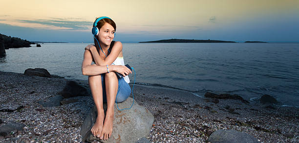 Earphones girl standing ocean shore enjoying sound closeup. Smiling beautiful woman listening headphones music on summer beach looking camera. Happy female traveler relaxing on rocky coastal nature.