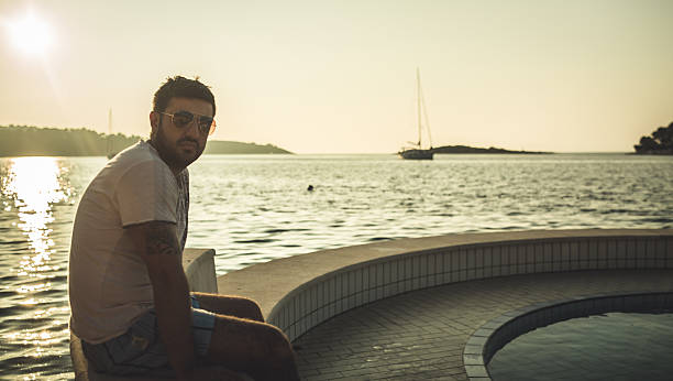 Handsome young man sitting on the pier/ dock at sunset Handsome good looking young man sitting on the pier/ dock at sunset with little yacht boat in the distance beautiful multi colored tranquil scene enjoyment stock pictures, royalty-free photos & images