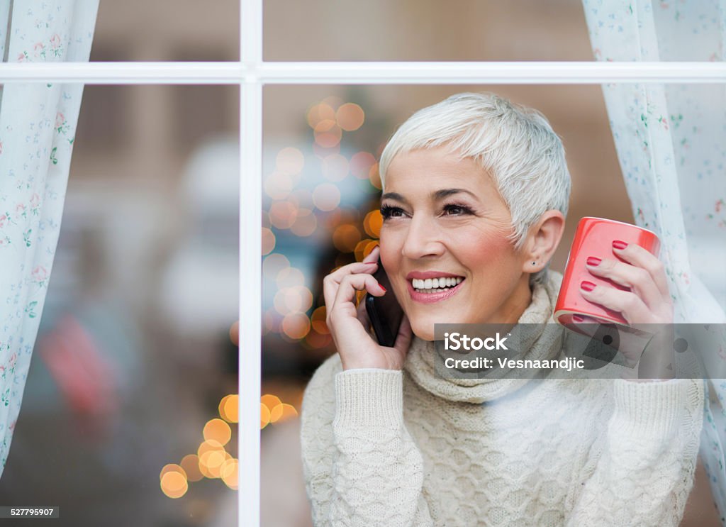 Beautiful smiling mature woman looking through window Beautiful smiling mature woman looking through window, enjoying in her warm home and coffee in winter time Winter Stock Photo