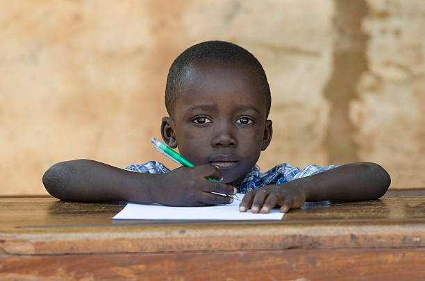 Sad Little African Boy At School Education Symbol Portrait of an African ethnicity schoolboy (age 4) in an educational environment in Bamako, Mali learning her lesson outdoors sitting on a desk and slightly smiling. sad african child drawings stock pictures, royalty-free photos & images