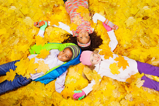 Three kids, girls and a boy laying on the ground covered with autumn maple orange leaves laying on the ground 