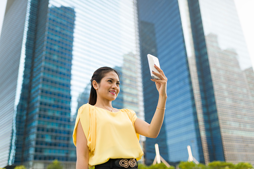 Attractive long hair Asian woman taking selfie with smart phone in business district, shot with wide angle lens using shallow depth of field  to blur out background details.