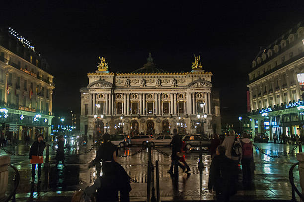 Place de l'Opera París, France - November 2, 2005: Place de l'Opera. Palais Garnier, home to the Opera House seen from the Place de l'Opera, a rainy November night. In the background, we see several cars stopped at a traffic light and, in the foreground, people walking on the sidewalk and others that leave or enter the subway station. place de lopera stock pictures, royalty-free photos & images