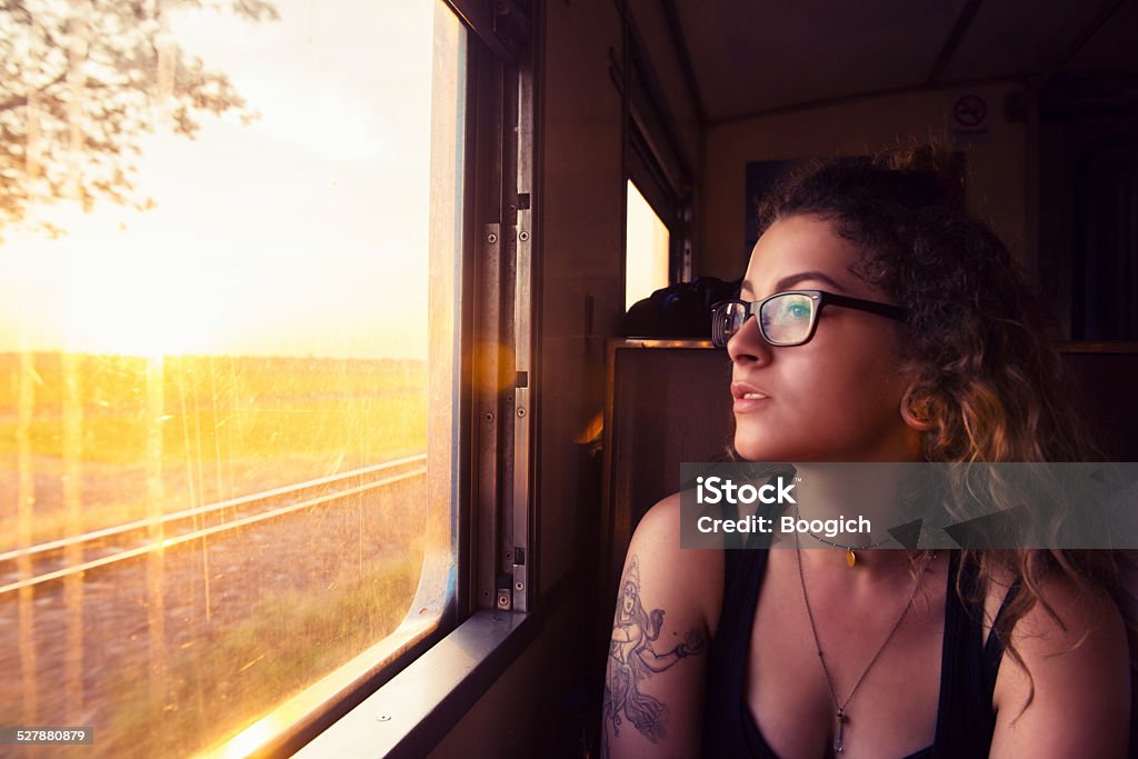 Young American Travels Thailand by Train at Sunrise As the morning sun rises over the countryside in Thailand a young American woman looks out the window of a train. The bright light casts a lens flare through the streaked glass window. She wears glasses and has curly blond hair. Gap Year Stock Photo
