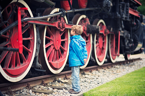 Little boy admiring a giant steam locomotive. The boy aged 5 is examining details of the red steel wheel that is bigger than the boy. The boy is wearing green jacket and jeans.
