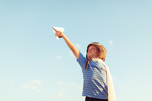 A young boy wearing a flight cap and aviator goggles holds up a paper airplane to the clear sky in hopes of flying. There is plenty of blue sky for copy space.