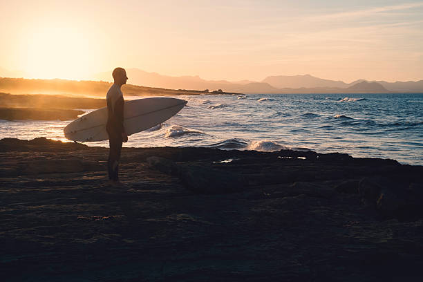 Young man surfer holding the surfboard while contemplating the waves at sunset.