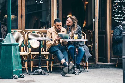 Young couple sitting in a french cafe outdoors and using digital tablet. Man is holding the tablet and they are both looking at it. They are wearing warm clothing.