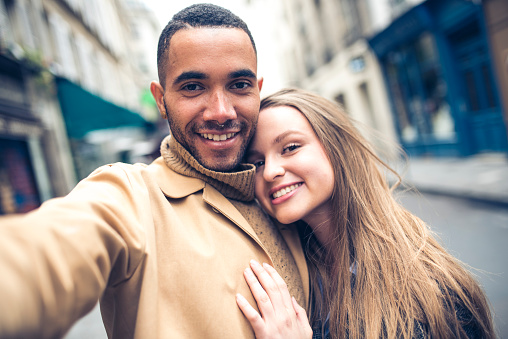 Young couple taking a selfie on city street in Paris, France. They are looking at camera, smiling.