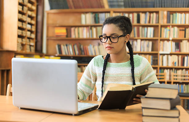 Concentrated young woman using a laptop in the library Attractive young woman with braids and eyeglasses, holding a book and working on laptop. Bookshelves as background. Stack of books on desk. bookstore book library store stock pictures, royalty-free photos & images