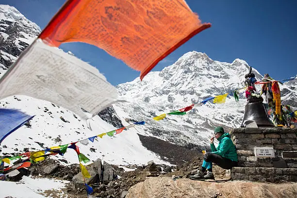 Yong woman mountaineer sitting and resting on the small stupa in memorial monument in Annapurna base camp, surrounded with amazing Annapurna mountain range on Himalayas. Praying flags are in foreground. Memorial is built in honour of climber who died in attempt to climb the summit. Annapurna is one of a fourteen mountains that is high more than 8000 metres above sea level. Weather is beautyful with clear sky