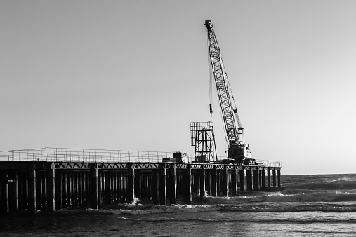 Construction on beach ocean sea pier jetty with crane and machinery in morning dawn silhouetted