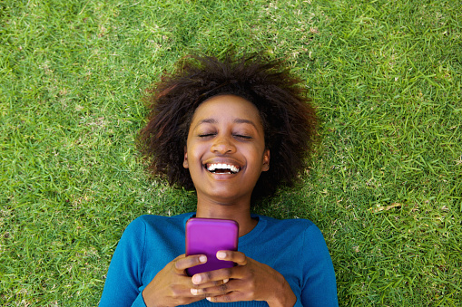 Portrait from above of a laughing woman lying on grass with cell phone