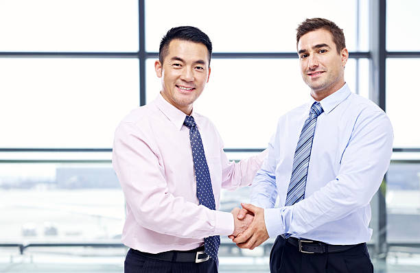 businessmen shaking hands at airport two businessmen, one asian and one caucasian, shaking hands looking at camera at modern airport. expatriate photos stock pictures, royalty-free photos & images