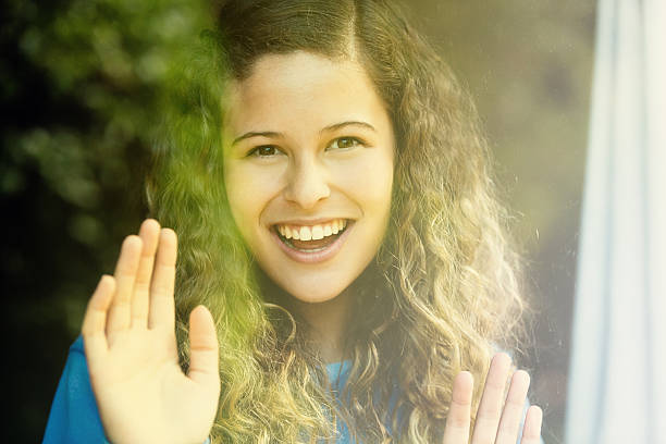 istock Beautiful young woman smiles and waves in greeting through window 528100809