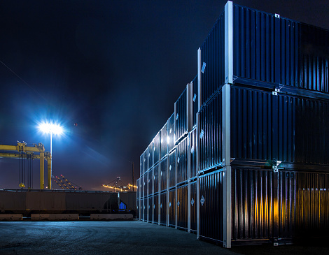 A blueish floodlight illuminates neatly stacked rows of shipping containers in an empty yard at the Port of Los Angeles/Long Beach