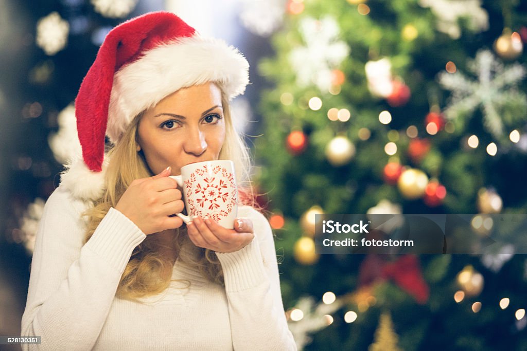 Woman drinking hot drink on Christmas eve Beautiful mid adult woman in Santa hat drinking hot drink next to the Christmas tree at home. 30-39 Years Stock Photo