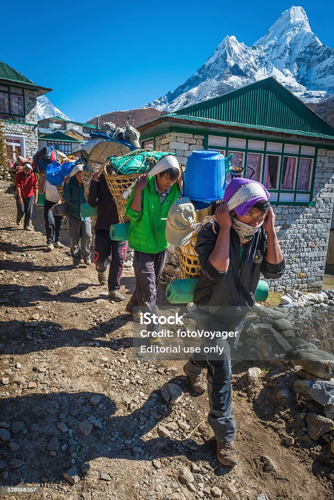 Line of Sherpa porters carrying expedition kit Himalaya mountains Nepal Namche Bazaar, Nepal - 17th October 2014: Line of Sherpa porters carrying heavy load with traditional headbands and wicker baskets along the Everest Base Camp trail high in the Khumbu valley below the iconic snowy spire of Ama Dablam (6812m) deep in the Himalaya mountain wilderness of the Sagarmatha National Park, Nepal, a UNESCO World Heritage Site. Guide - Occupation Stock Photo