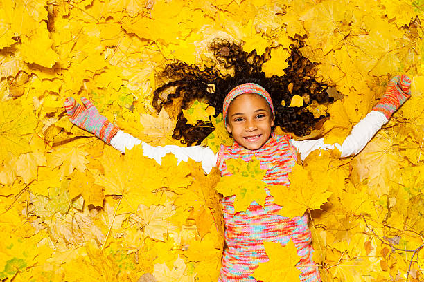 Smiling African little girl laying on the yellow background and covered with autumn maple leaves 
