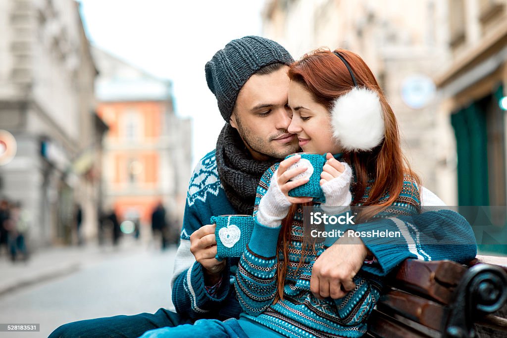 Young couple with coffee in winter outside Young loving couple dressed in blue sweater sitting with knitted coffee cups on the bench at the old city in winter Couple - Relationship Stock Photo