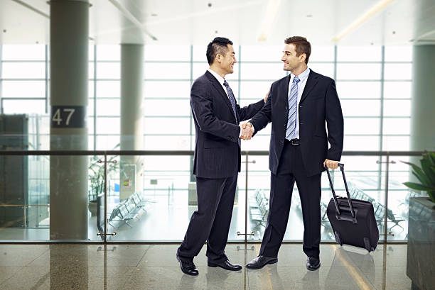 business people shaking hands at airport two businessmen, one asian and one caucasian, shaking hands and smiling at modern airport. expatriate photos stock pictures, royalty-free photos & images