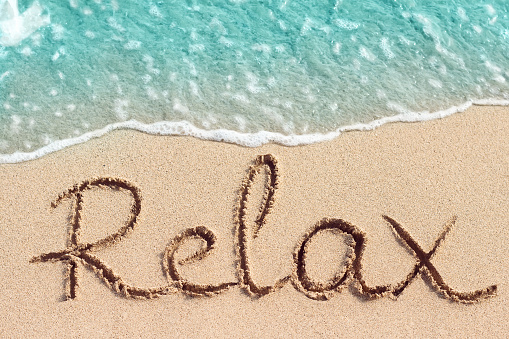Word Relax handwritten on sandy beach with blue wave on background