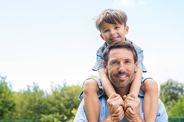 Father giving piggyback ride Happy father giving shoulder ride on his shoulders in garden. Happy smiling boy on shoulder dad looking at camera. Father and son enjoying outdoor. on shoulders stock pictures, royalty-free photos & images