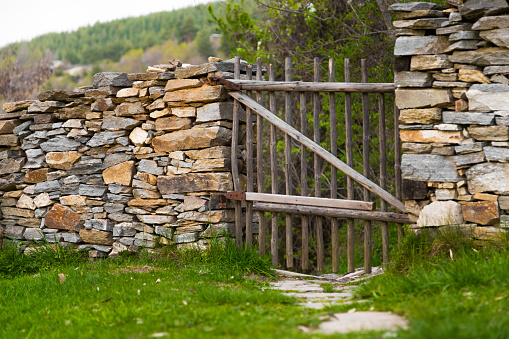 Rustic handmade wooden gate made from natural wooden poles in a stone wall with a path leading to a rural hillside.