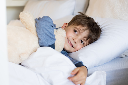 Young boy sleeping on bed. Cute little boy lying in bed and smiling in the morning time. Small boy hugging his teddy bear for his afternoon nap.