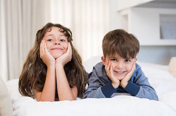 Children lying on front on bed. Cute little girl and boy lying on white bed and looking at camera. Happy smiling brother and sister in a playful mood after waking up in the morning.