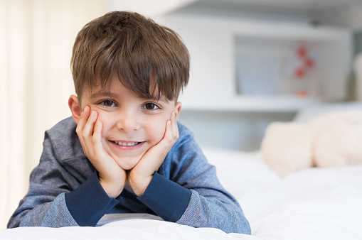 Handsome boy with toothy smile looking at camera. Lovely child lying on front in bed. Happy young cute boy lying on bed.