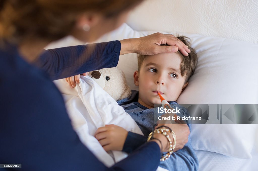 Boy measuring fever Sick boy with thermometer laying in bed and mother hand taking temperature. Mother checking temperature of her sick son who has thermometer in his mouth. Sick child with fever and illness while resting in bed. Child Stock Photo