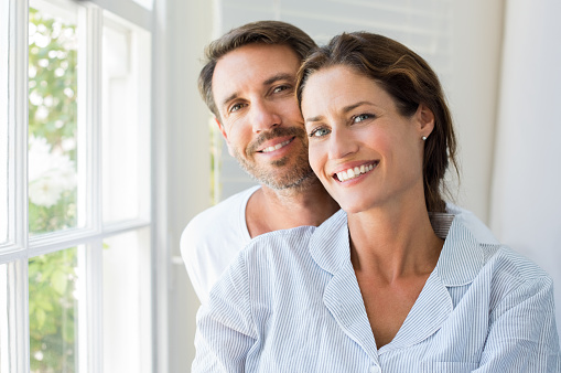 Happy young couple sitting near window in the bedroom. Portrait of smiling man and woman looking at camera at home. Couple in pajamas during the morning.