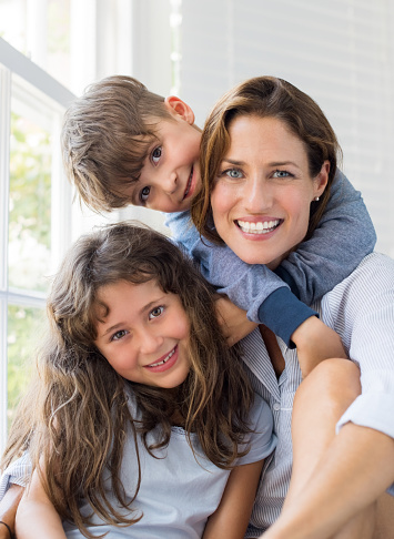Son and daughter embracing mother. Mother enjoying with children on a bright sunny morning. Portrait of happy smiling mom with her children at home looking at camera. Happy family embracing.
