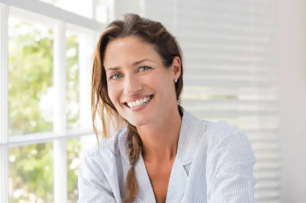 Portrait of beautiful mature woman sitting and smiling. Woman feeling fresh on a bright sunny morning at home. Woman sitting in nightwear near the window and looking at camera.