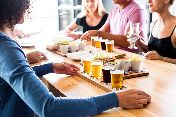 Group Of Friends Enjoying Lunch & Craft Beer Tasting Group of friends seated at a table inside a brewery at daytime enjoying lunch & craft beer tasting together. craft beer stock pictures, royalty-free photos & images