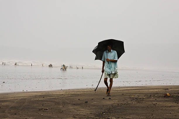 Ratnagiri, MH, India - August 27, 2011: Taken this picture while on my trip to Ratnagiri beach during monsoon season. Tried to capture this old man in foreground walking on the beach while it was raining and windy. In background are the tourists playing and enjoying on the beach.