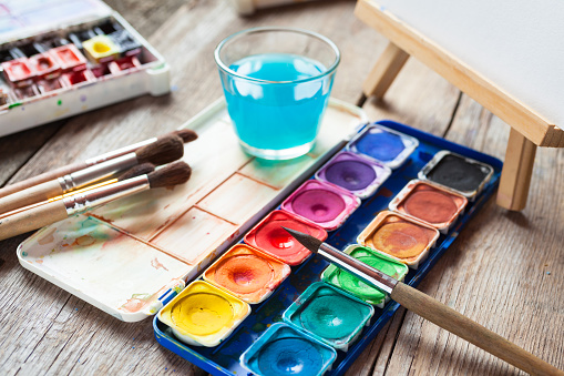 Set of watercolor paints, art brushes, glass of water and easel with painting on old wooden table.