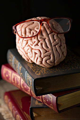 An extreme close up vertical  photograph of a replica of a human brain wearing eye glasses and sitting on a stack of fine literature books. 