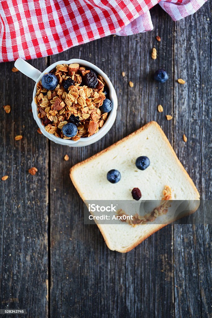 Healthy and funny breakfast: granola and white toast bread Healthy and funny breakfast for kids: homemade golden crunchy granola with almonds and raisins and white toast bread with fresh blueberries and granola smiling face, wooden table background Animal Stock Photo