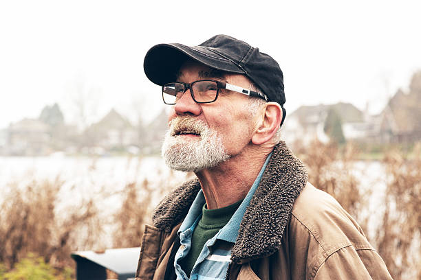 Senior man with beard wearing glasses and black cap outdoor in winter landscape with houses in the background.