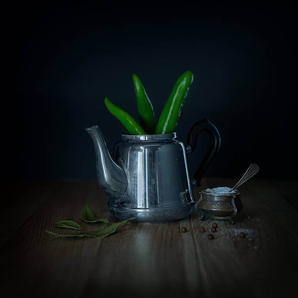 Green chili peppers in a metal teapot, laurel leaves and salt on the wooden table. Rustic style. Defocused black background