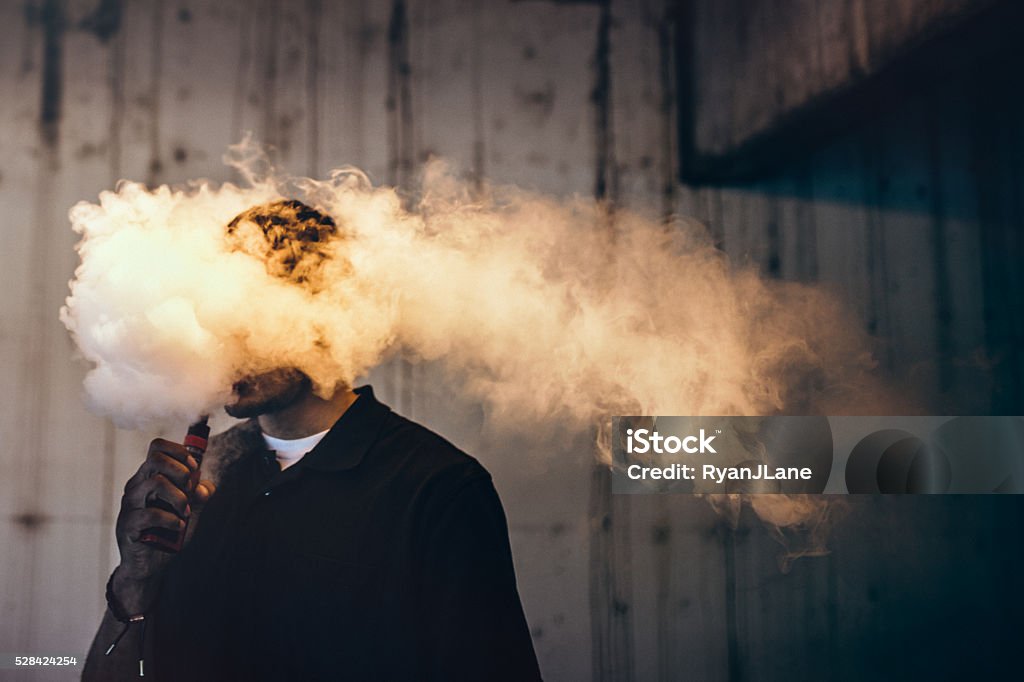 Man Using An Electric Cigarette A dark and high contrast image of a young adult male has a cloud of nicotine vapor "smoke" pouring from his mouth.  Vape smoking, or "vaping" is growing in popularity, as well as falling under stricter state and governmental regulations.  Horizontal image with copy space. Electronic Cigarette Stock Photo