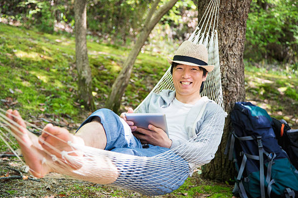 Portrait of a mature man relaxing in a hammock Portrait of a mature man looking at the camera whilst relaxing outdoors in a hammock and holding a digital tablet. Okayama, Japan. April 2016 hammock men lying down digital tablet stock pictures, royalty-free photos & images