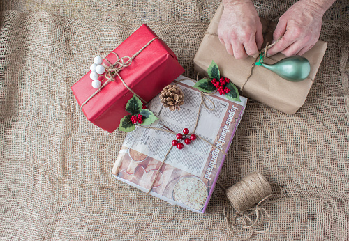 very rustic country christmas image with man's hands wrapping christmas gift with brown paper and tying with baler twine with another gift wrapped in news paper with ginger bread story on it lying on a burlap background