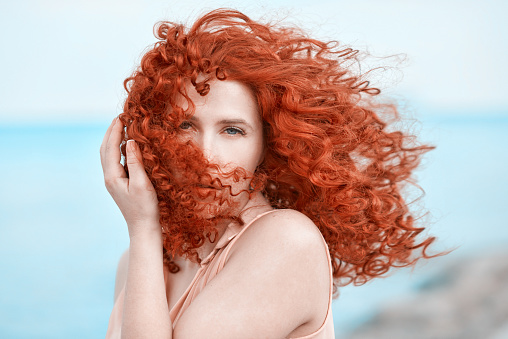 beautiful red and curly hair woman touching hair and posing on the beach, looking at camera.