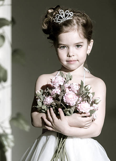 Lovely little girl in small crown dressed a white ball gown. The little girl is holding flowers in hands. She is looking at the camera. Studio shooting